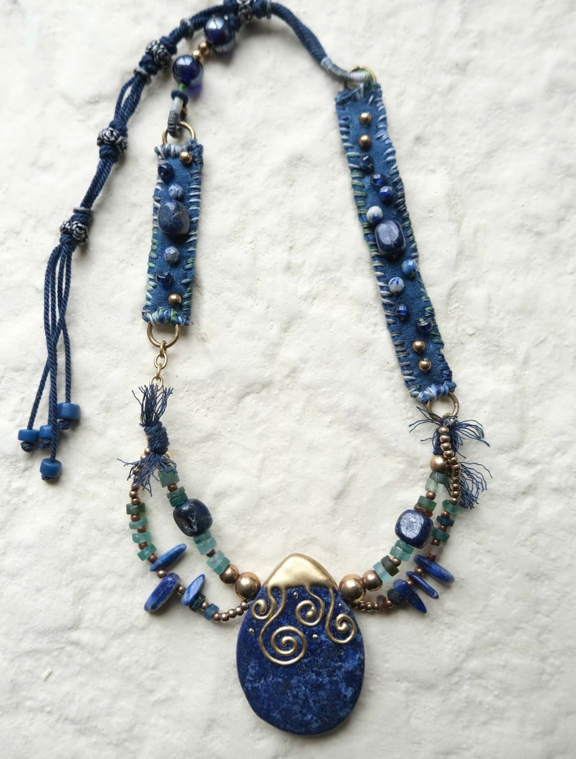 Flat lay of a handmade blue stone pendant necklace with stitched textile panels, mixed beads, and metal accents.